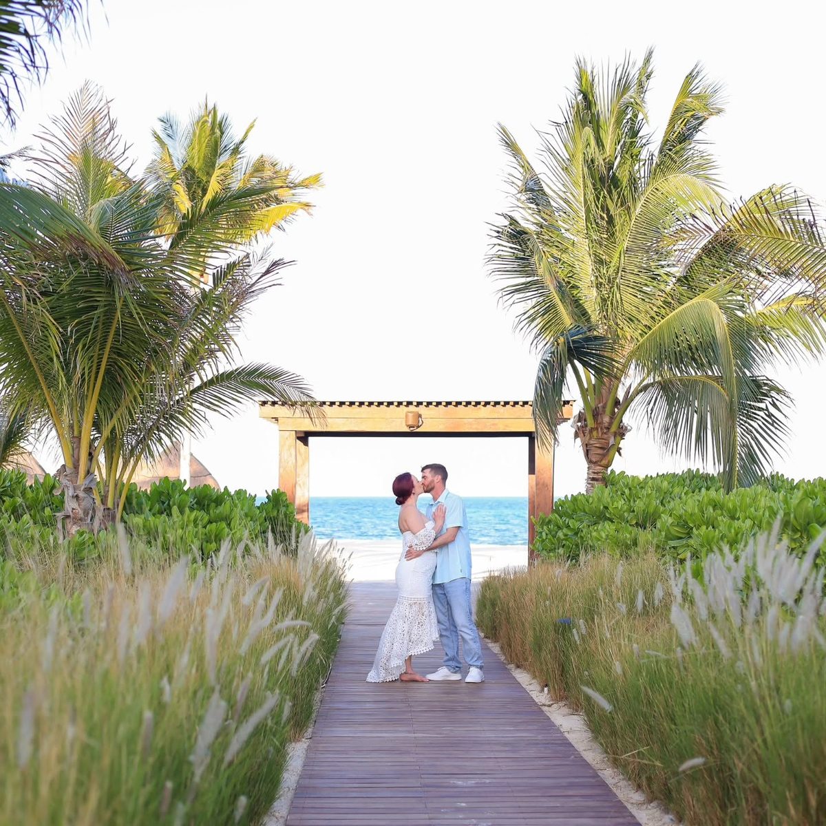Jackie and Jered kissing on a boardwalk framed by palm trees with the ocean behind